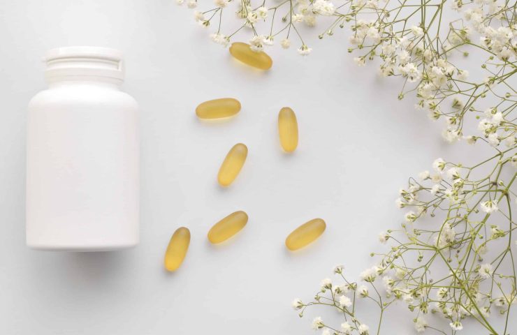 Oil yellow gelatin capsules with white bottle on white background with flowers, vitamins and antioxidant concept Omega 3, liver cod or evening primose oil for healthcare. Minimalism. Copy space.